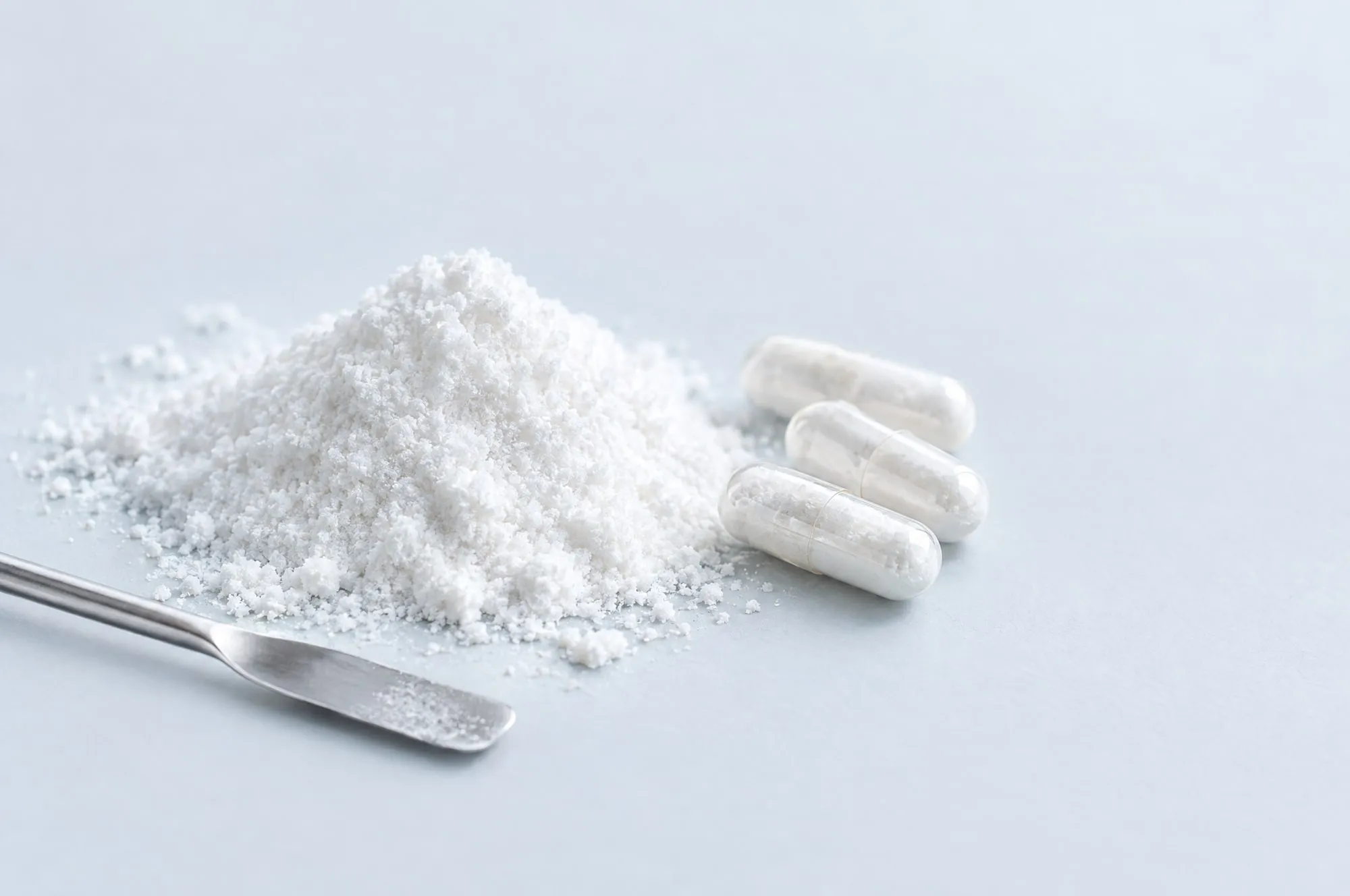 A mound of white citicoline powder beside clear capsules and a steel spatula