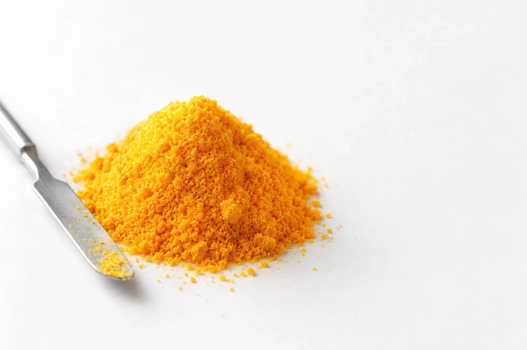 A mound of fine orange-yellow riboflavin powder beside a steel laboratory spatula